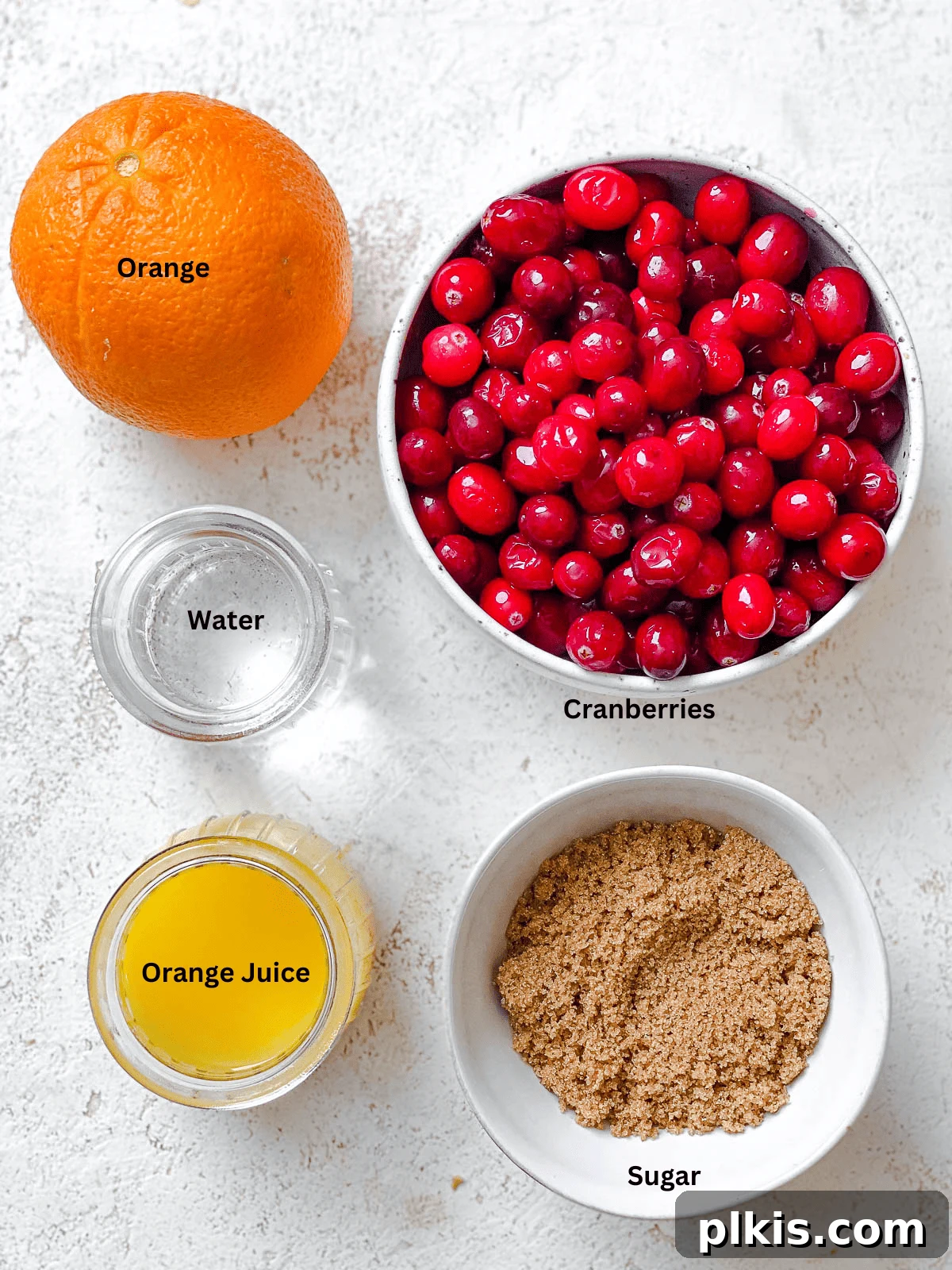 A selection of ingredients for Easy Cranberry Sauce with Orange Juice, including fresh cranberries, oranges, sugar, and a small pot, arranged on a white surface.