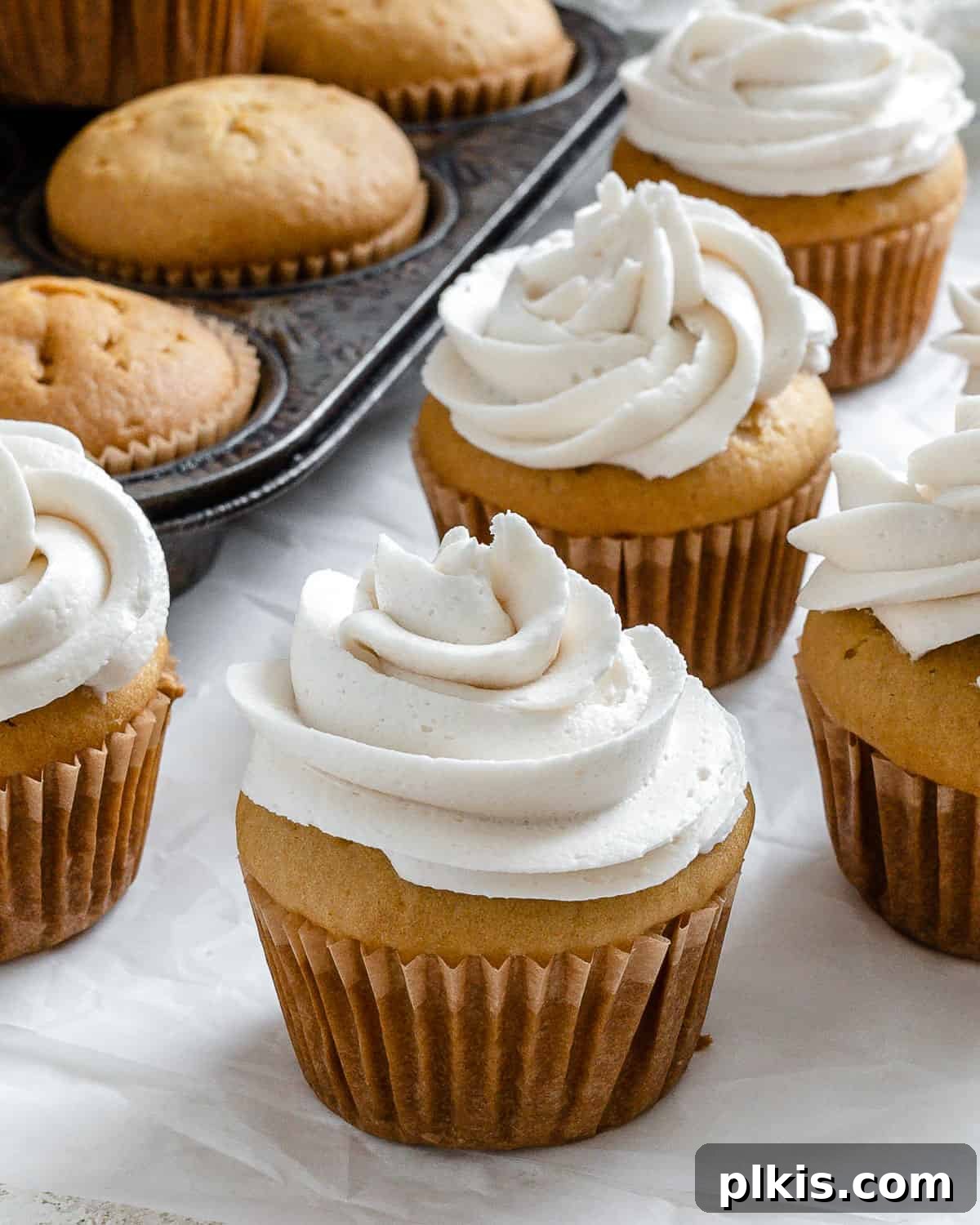 Effortless Vegan Vanilla Cupcakes 2 completed vegan vanilla cupcakes spread out on white surface and in muffin tin, showcasing their fluffy texture and golden tops