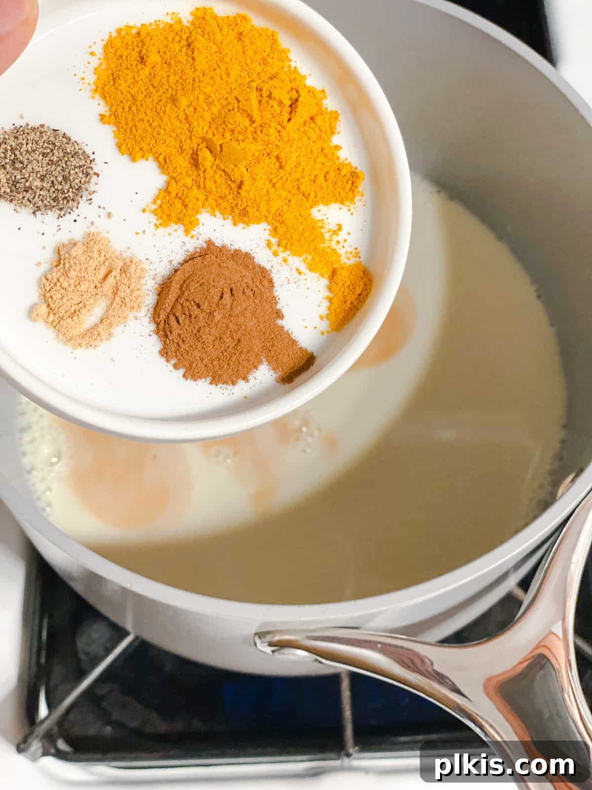 Golden Turmeric Elixir 7 Process shot showing a blend of turmeric, cinnamon, and ginger spices being added to the simmering milk in a saucepan.