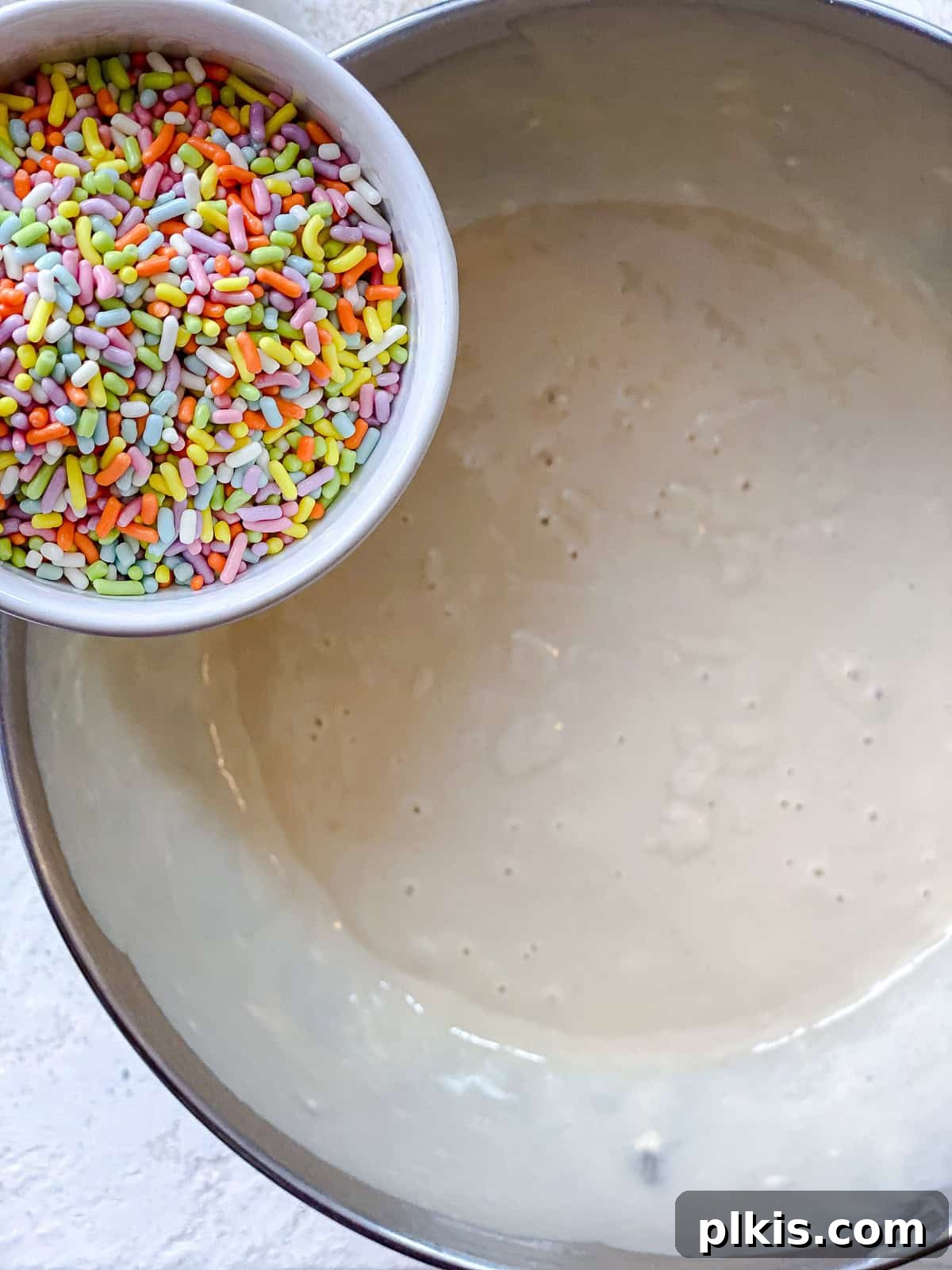 Rainbow Vegan Cupcake Magic 4 Process shot showing vibrant rainbow sprinkles being carefully folded into a light-colored cupcake batter in a mixing bowl.