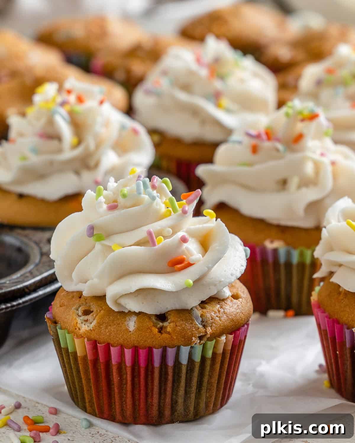 Rainbow Vegan Cupcake Magic 6 A close-up shot of several completed Vegan Funfetti Cupcakes, beautifully frosted and adorned with sprinkles, placed on a white surface alongside a cupcake tin.