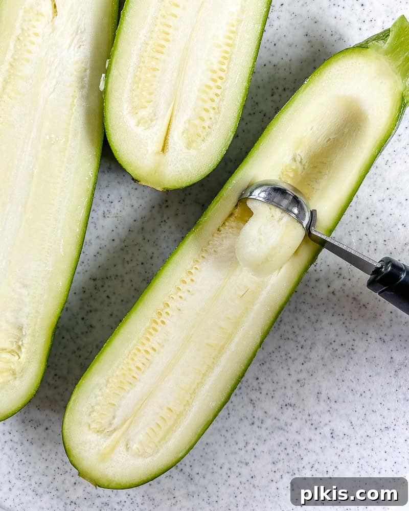 Process shot illustrating how to scoop out the seeds and flesh from the center of a zucchini half with a spoon, creating a boat shape.