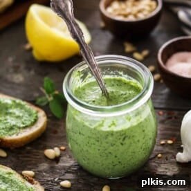 A completed batch of fresh zucchini pesto in a clear glass jar, resting on a brown surface with scattered ingredients in the background.
