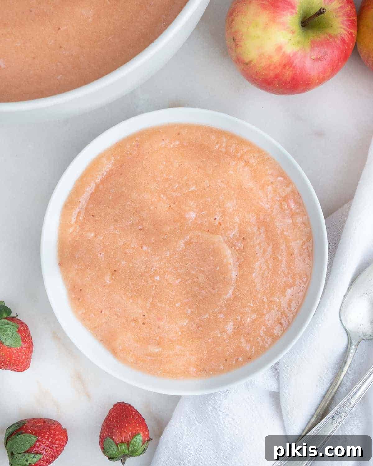 48 Irresistible Vegan Apple Delights 45 Completed Strawberry Apple Sauce in a white bowl against a white background with apples and strawberries in the background.