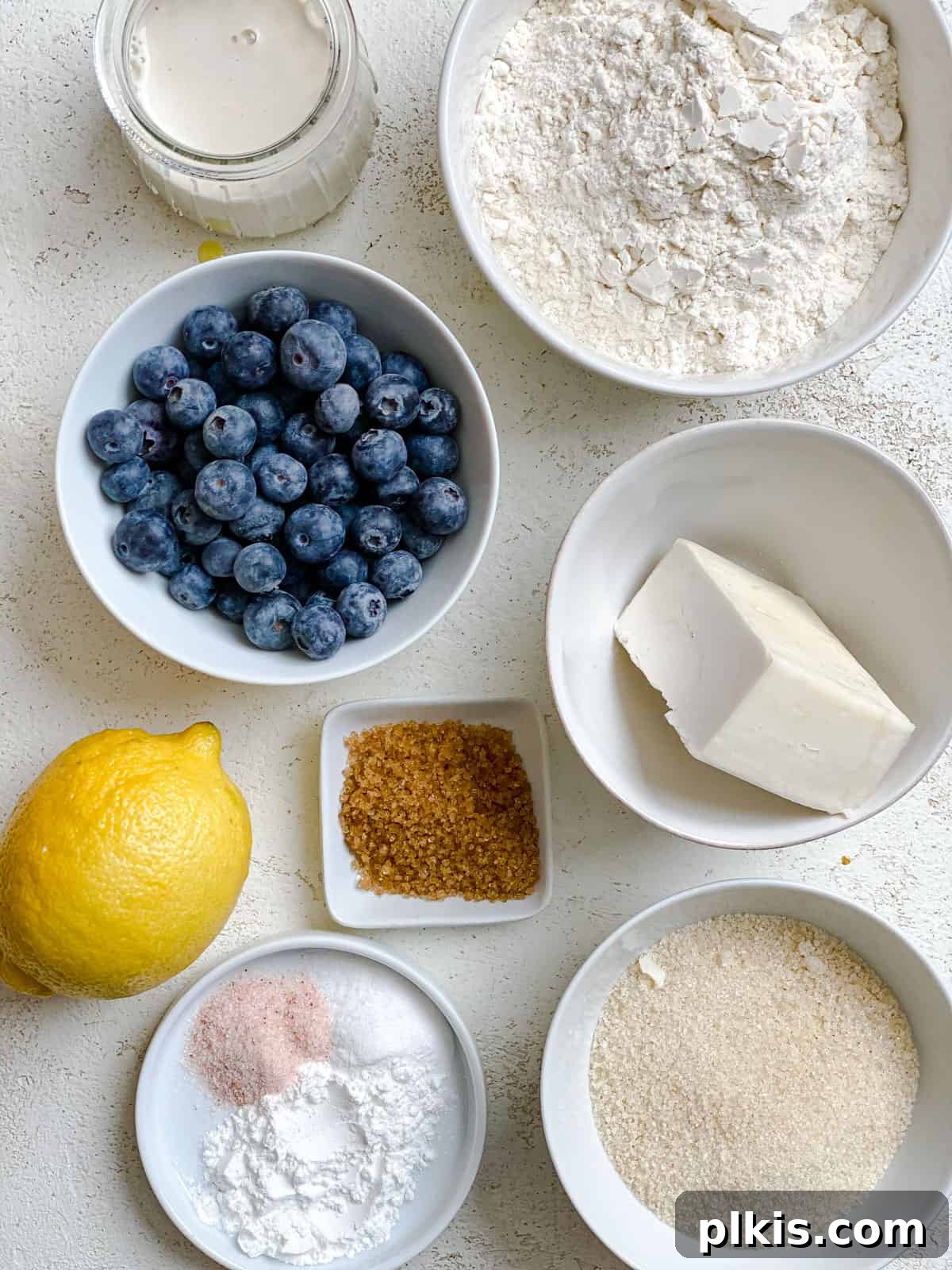 Sunshine Lemon Blueberry Plant-Based Scones 3 Ingredients for Vegan Lemon Blueberry Scones measured out against a white surface, including flour, sugar, butter, lemons, and blueberries.