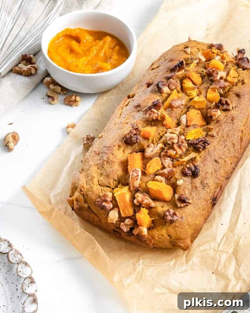 Finished vegan pumpkin bread loaf on parchment paper, with ingredients like pumpkin and spices blurred in the background.