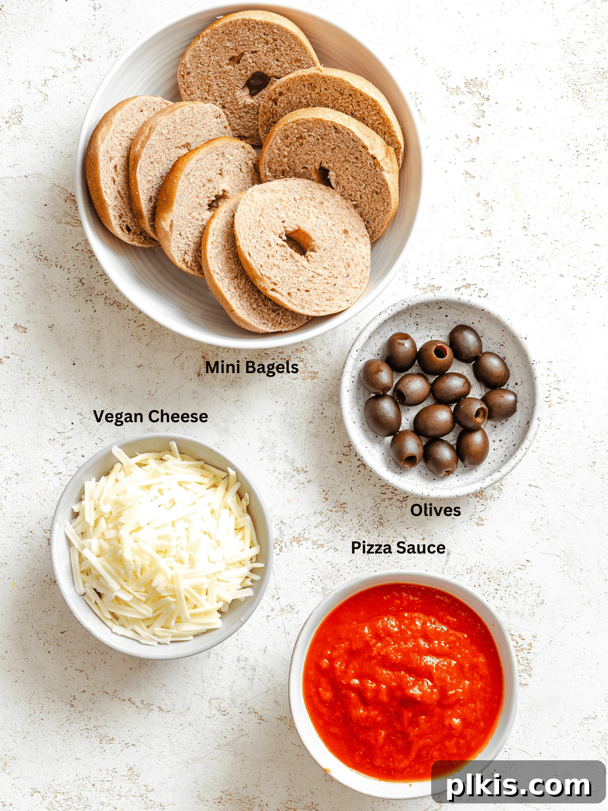 Spooky Spider Pizza Bites 3 All the core ingredients for Mini Spider Pizza Bagel Bites, including mini bagels, pizza sauce, vegan cheese, and black olives, neatly measured out on a white surface.