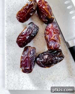 Process image showing six whole Medjool dates on a white cutting board next to a knife.