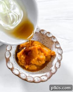Process image showing wet ingredients like vanilla extract or maple syrup being poured into a bowl of pumpkin puree.