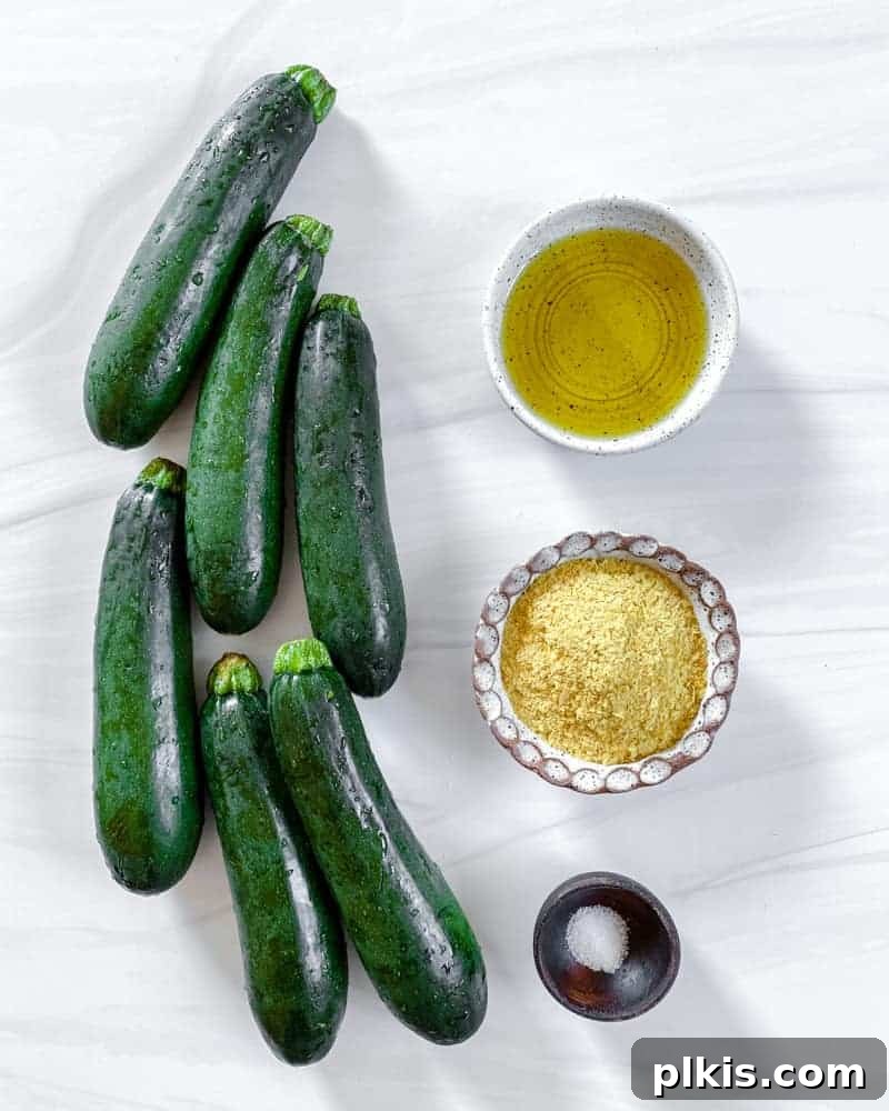 Crispy Vegan Cheesy Zucchini Bites 3 A selection of fresh whole zucchini and various ingredients like olive oil, nutritional yeast, and salt in bowls, arranged for making vegan cheesy zucchini chips against a clean white background