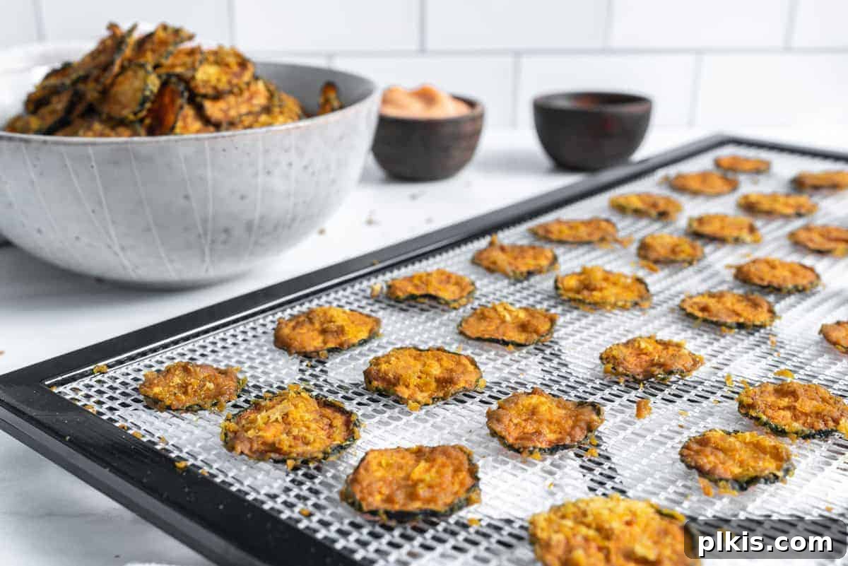 Crispy Vegan Cheesy Zucchini Bites 5 Cooked cheesy zucchini chips carefully arranged on a dehydrator rack, with a full bowl of chips in the blurred background, showcasing the delicious end product