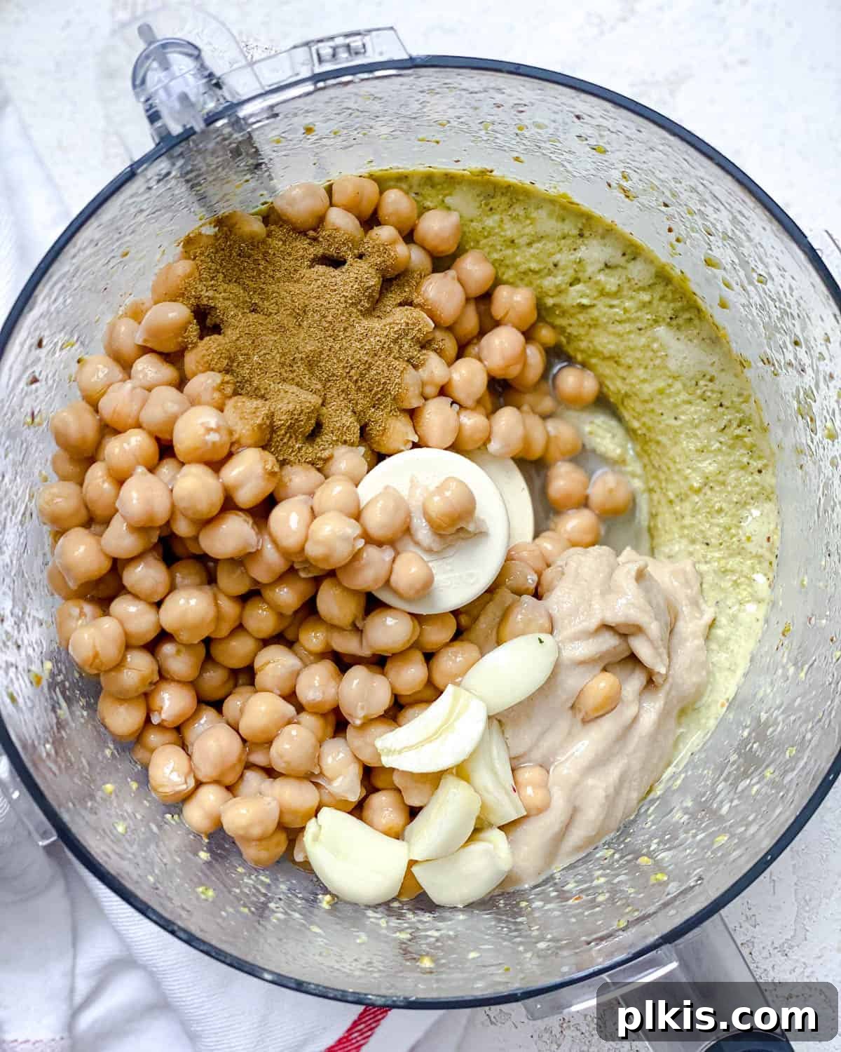 Emerald Pistachio Hummus 4 Process shot: All remaining ingredients, including chickpeas, lemon juice, garlic, cumin, tahini, and ice, being added to the food processor.
