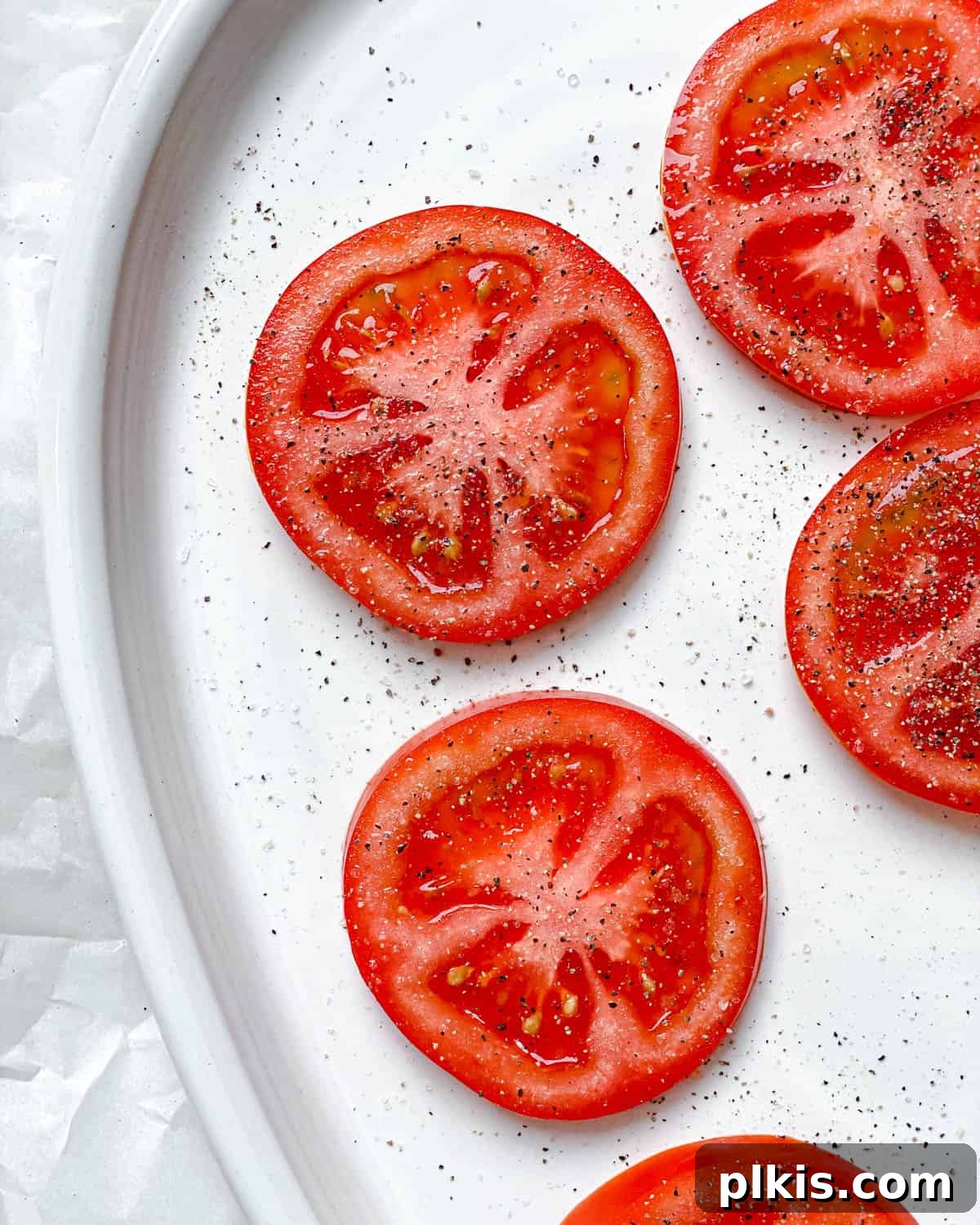 Plant-Based Caprese Delight 4 Process shot showing perfectly sliced ripe tomatoes on a white plate, seasoned with salt and pepper.