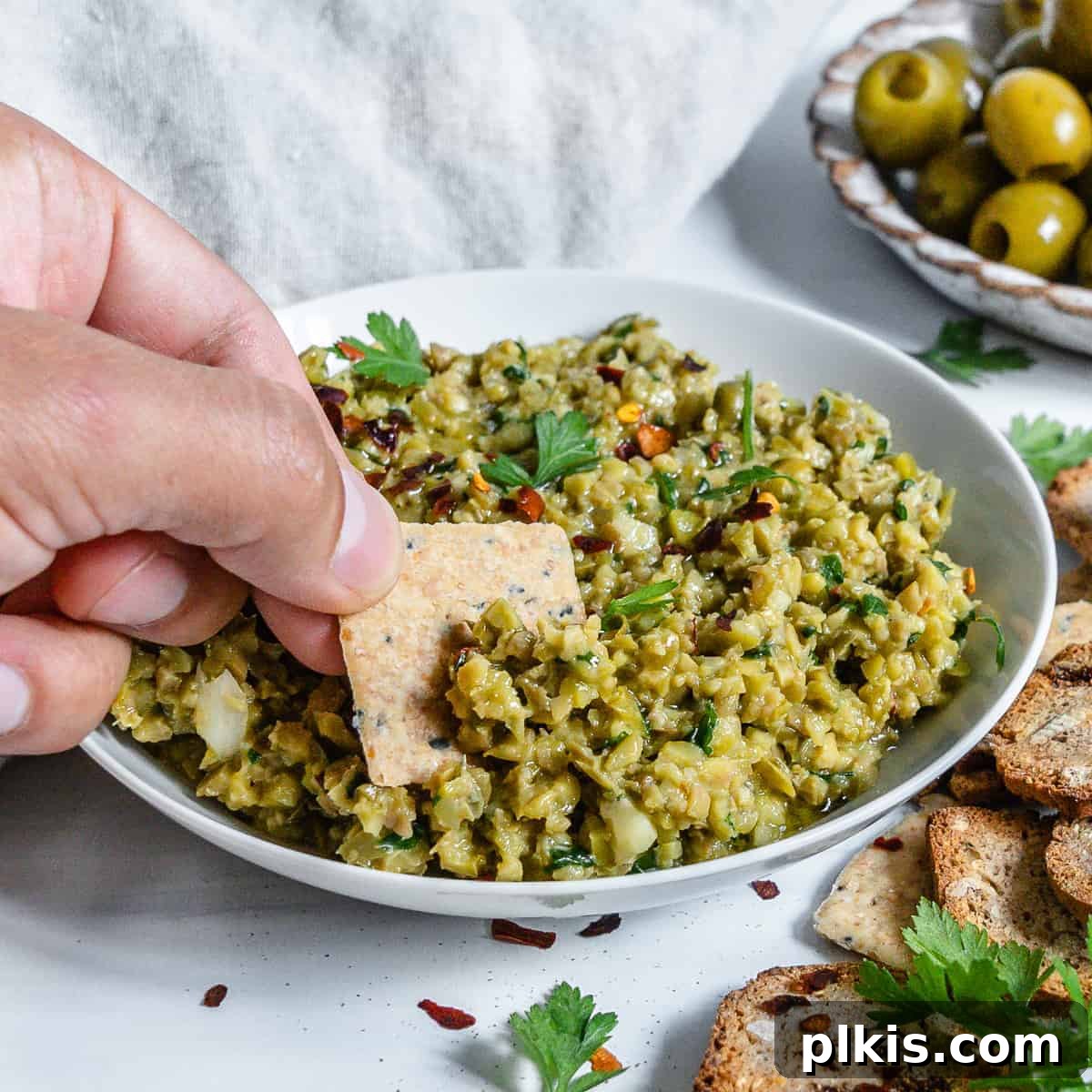 Vibrant Green Olive Spread 2 A bowl of vibrant green olive tapenade, with a cracker being dipped into it. The background features fresh ingredients and olives, suggesting homemade goodness.