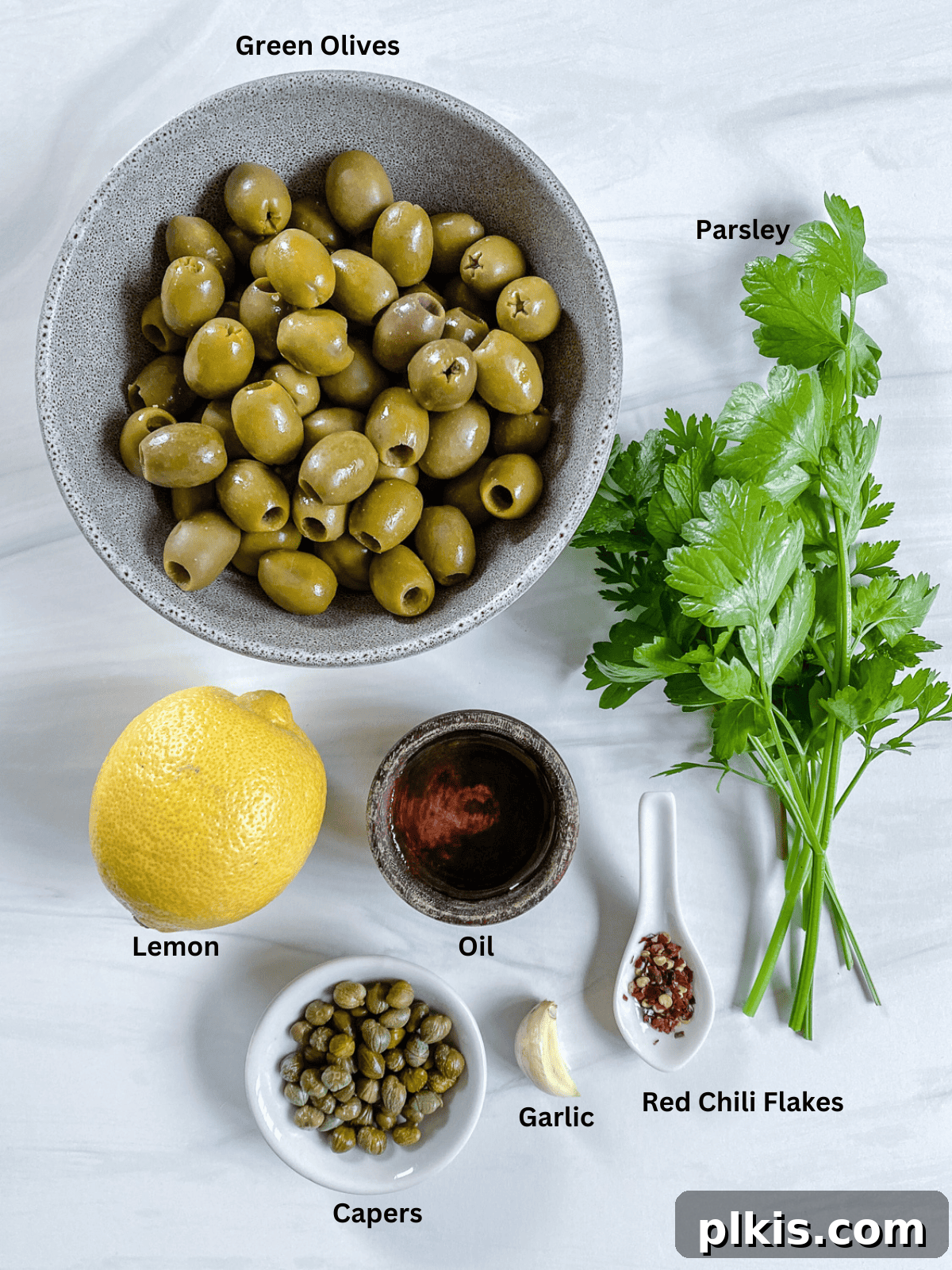 Vibrant Green Olive Spread 3 All the fresh ingredients for Green Olive Tapenade, including green olives, capers, garlic, and parsley, are neatly laid out on a clean white surface, ready for preparation.