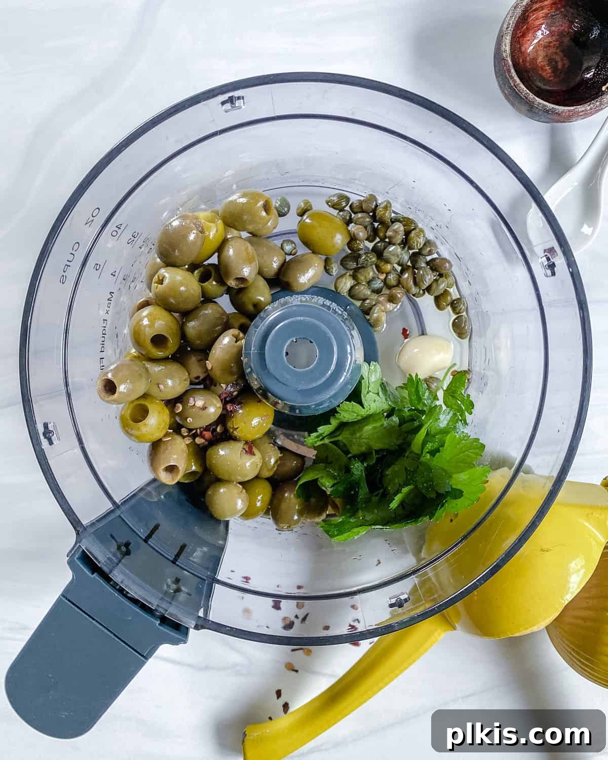Vibrant Green Olive Spread 4 Green olives, capers, and garlic cloves being added into the bowl of a food processor against a clean white background, ready to be blended into tapenade.