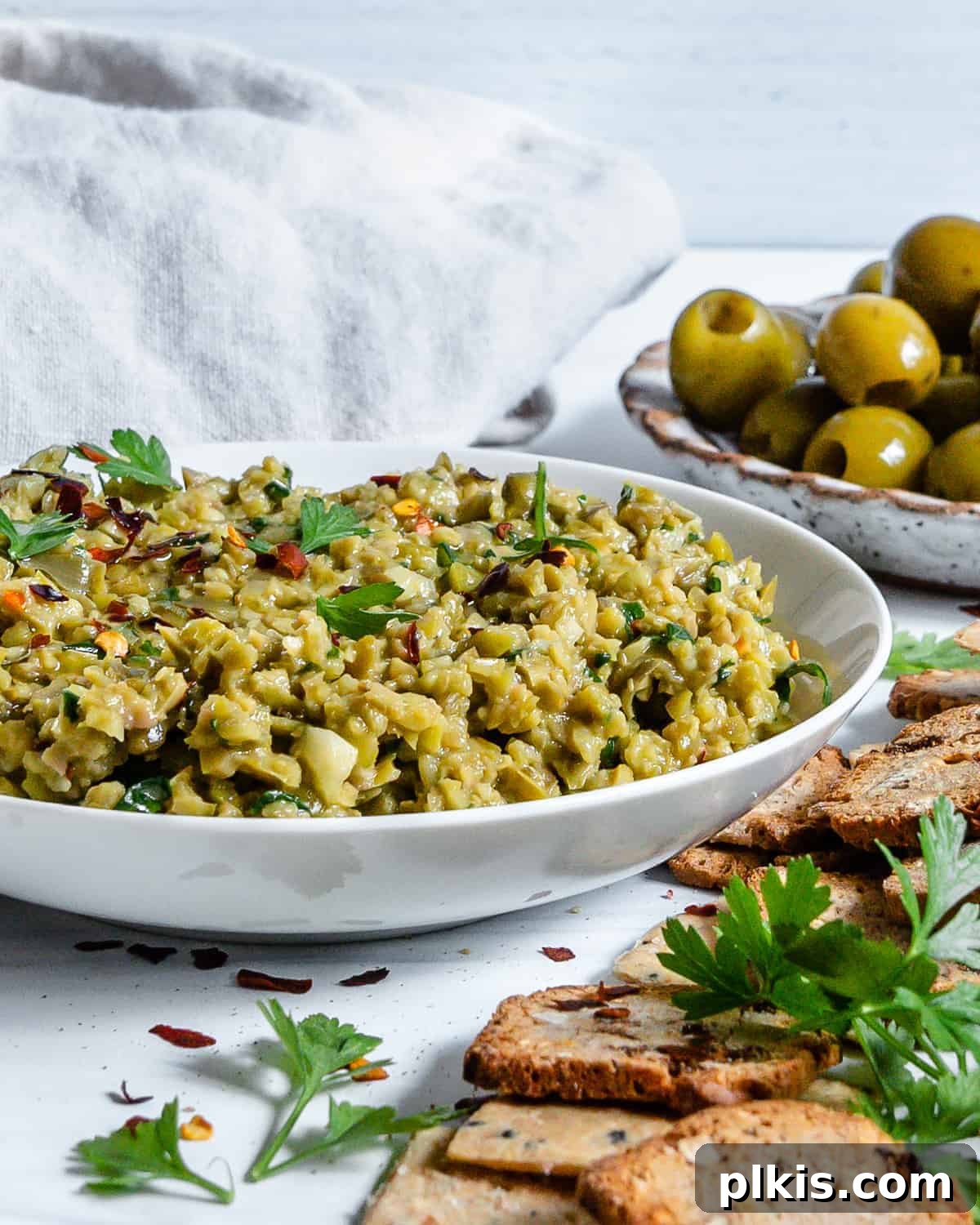 Vibrant Green Olive Spread 5 A beautiful white bowl filled with freshly made Green Olive Tapenade, accompanied by a single cracker poised for dipping. The background is softly blurred, highlighting the vibrant texture of the spread.