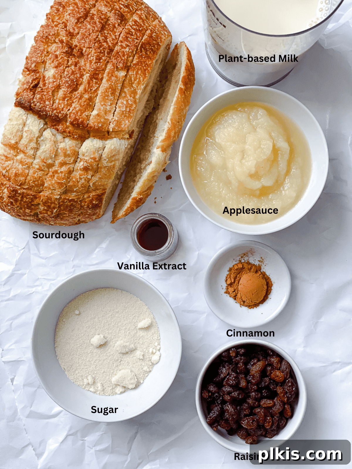 Decadent Vegan Bread Pudding 3 Assortment of ingredients for Vegan Bread Pudding, including bread, plant-based milk, applesauce, sugar, cinnamon, vanilla extract, and raisins, neatly arranged on a white surface.