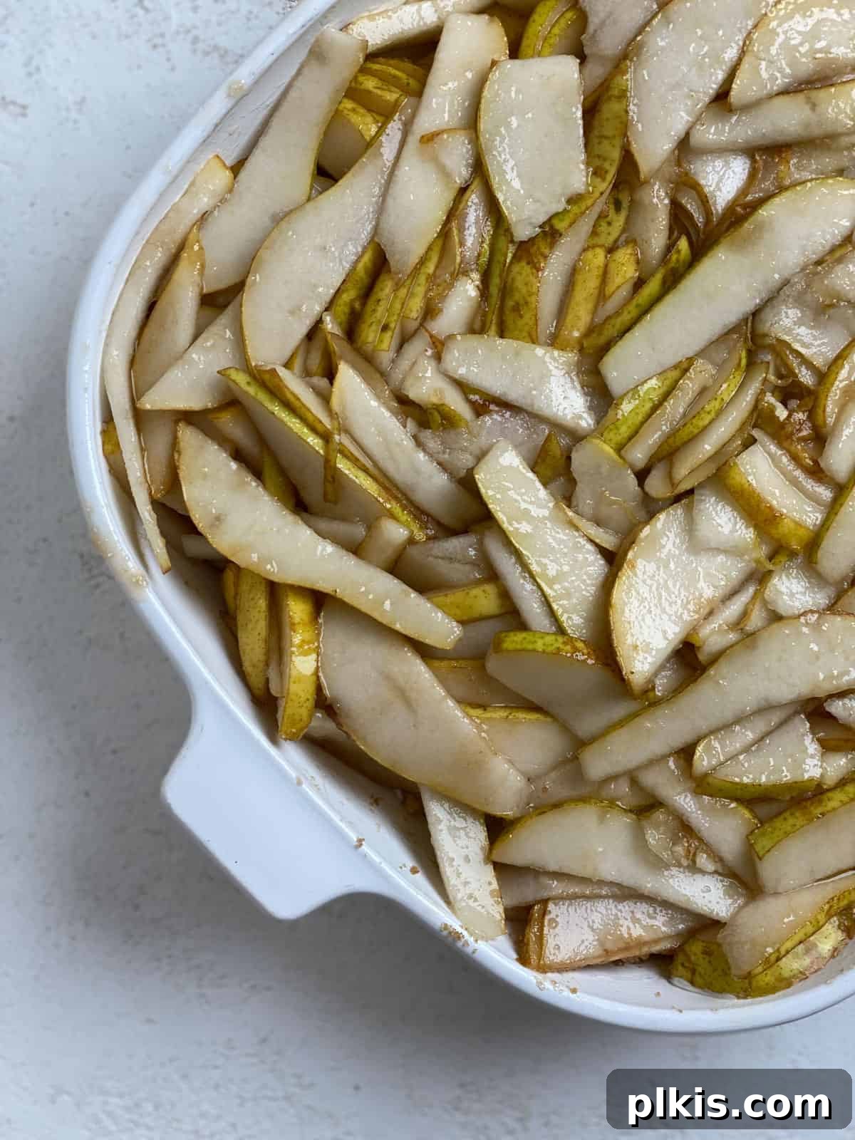 Process shot showing thinly sliced pears mixed with a quarter of the brown sugar in a prepared baking dish, forming the base of the Easy Pear Crisp.