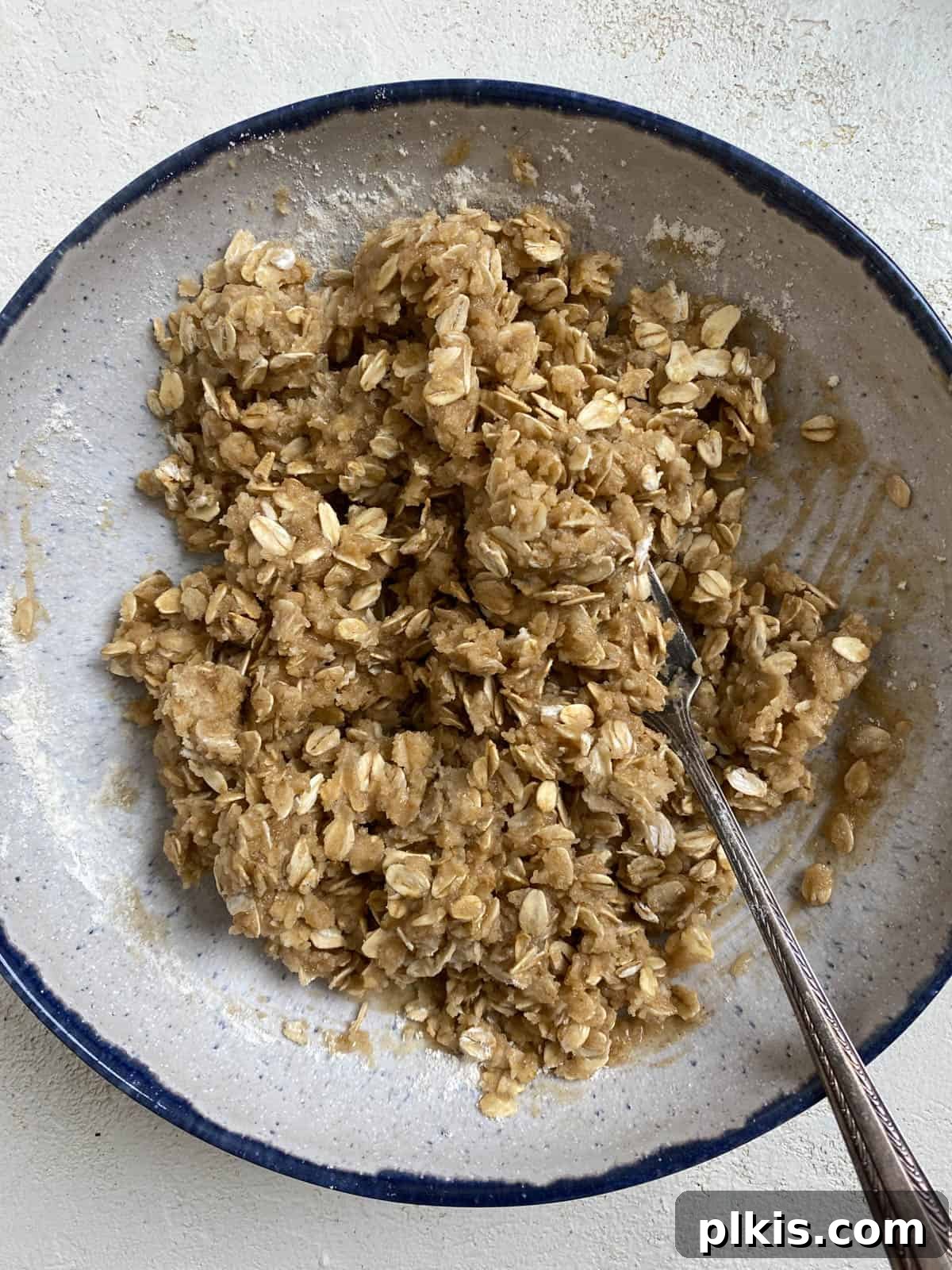Process shot showing the dry and wet ingredients for the streusel topping (flour, oats, sugar, spices, vanilla, and melted vegan butter) being combined in a separate bowl to achieve a crumbly texture.