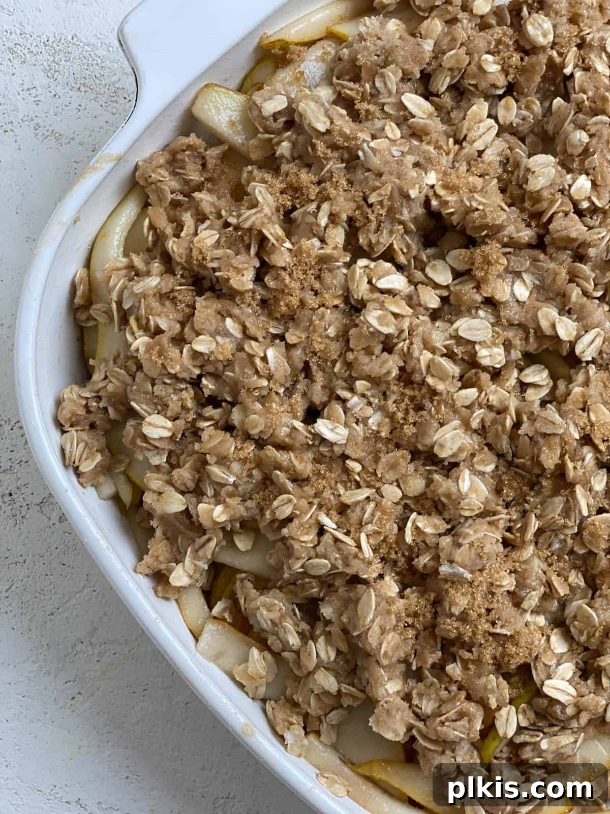 Process shot showing the golden, oat-rich streusel topping being evenly distributed over the sweet pear mixture in the baking dish, creating the iconic layers of the Easy Pear Crisp.