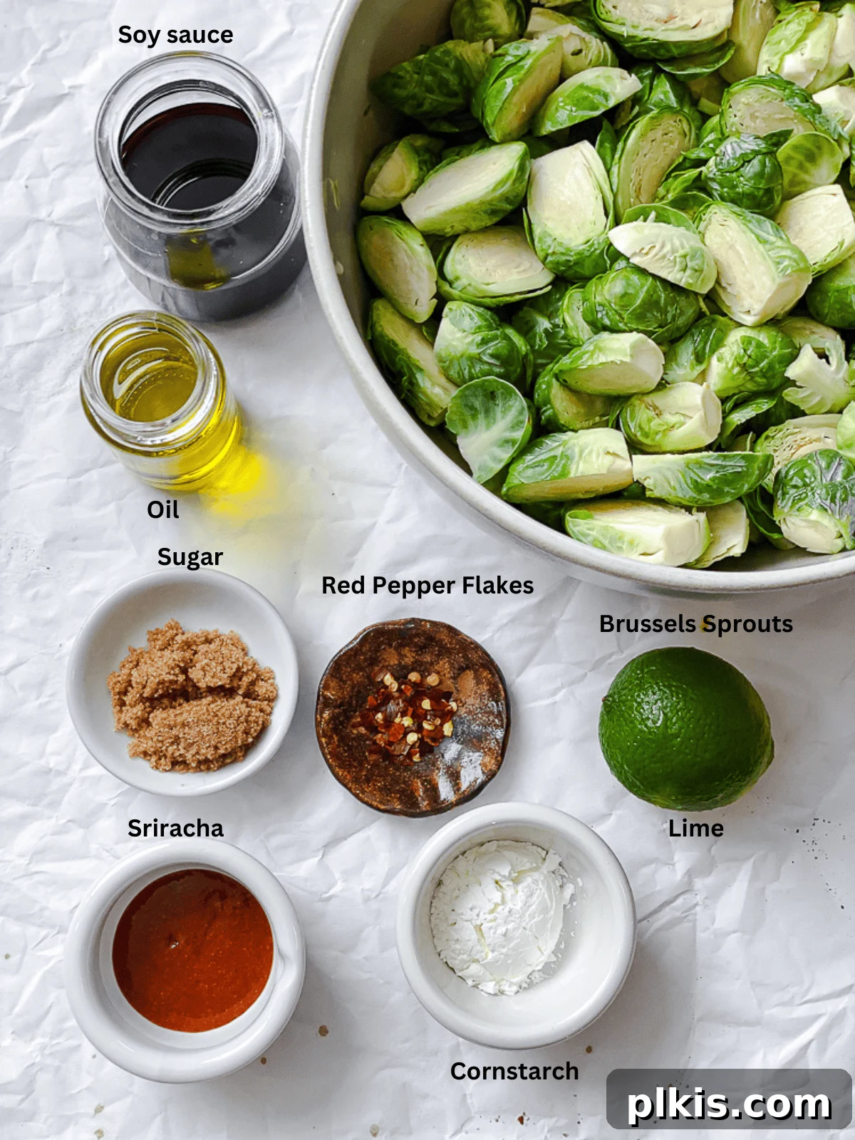 Ingredients for Crispy Brussels Sprouts measured out on a white surface, including soy sauce, sriracha, lime, and fresh sprouts.