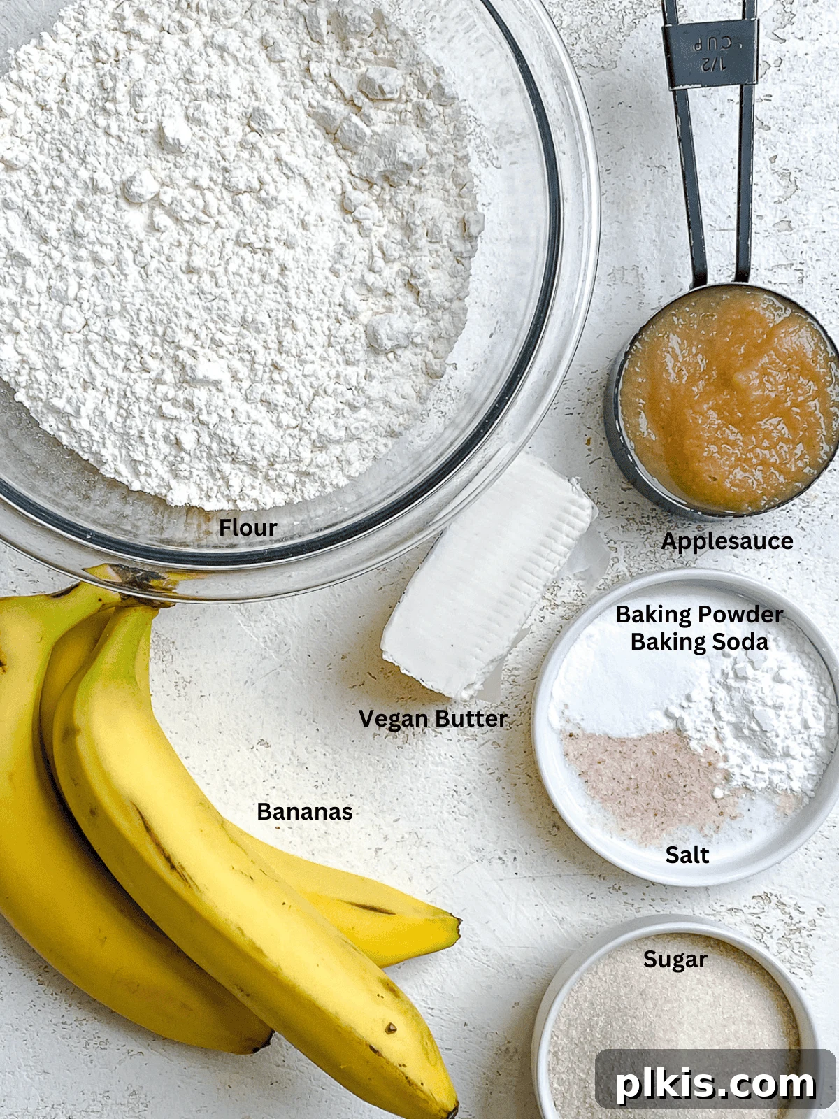 Effortless Vegan Banana Bread Applesauce Enhanced 3 A curated display of ingredients for Easy Vegan Banana Bread (With Applesauce) neatly arranged on a pristine white surface, highlighting pantry staples like bananas, flour, sugar, and applesauce.