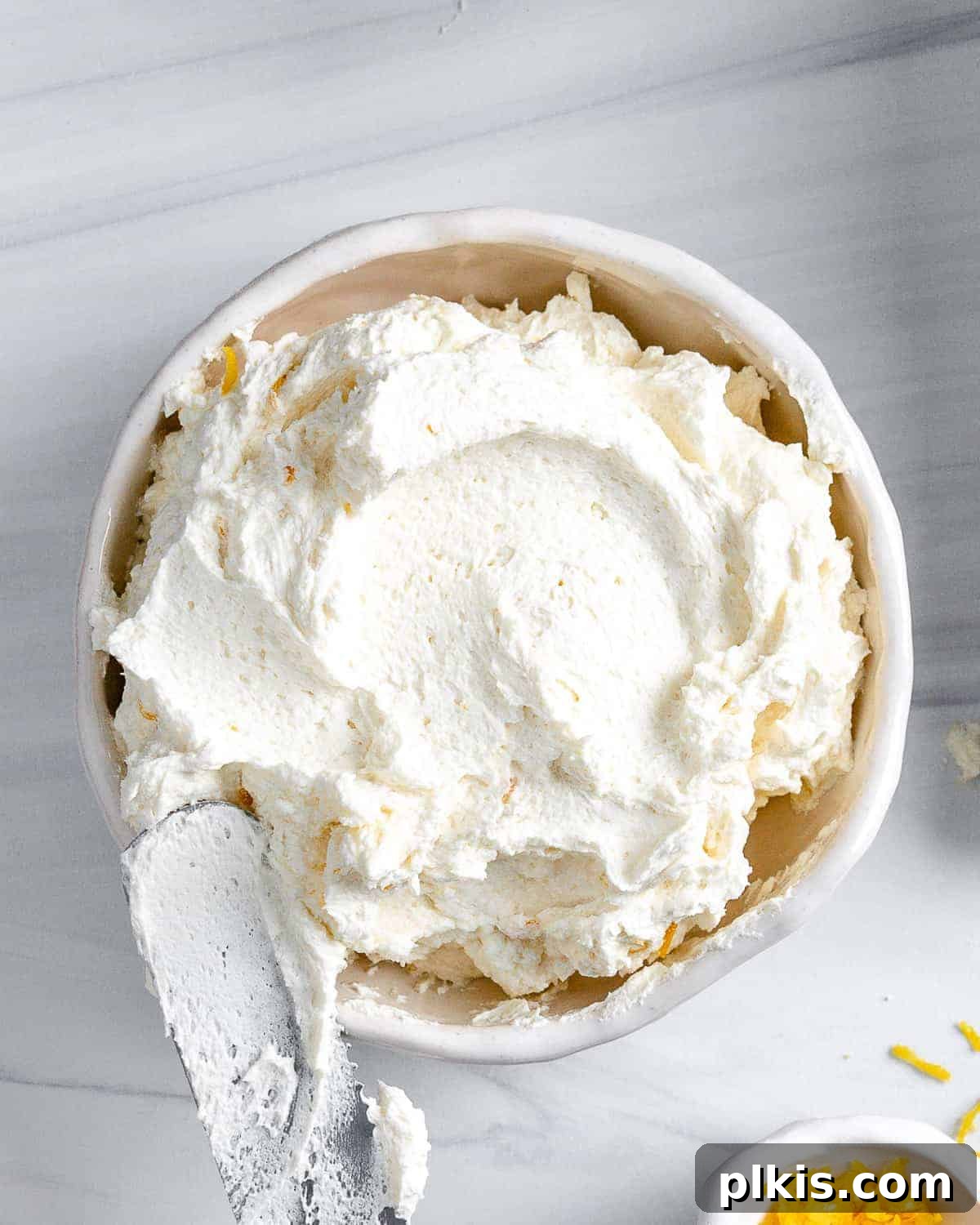 Luscious Vegan Lemon Buttercream 2 A perfectly prepared bowl of light and fluffy vegan lemon buttercream frosting, ready to be enjoyed. The creamy texture and vibrant yellow hue are inviting, set against a pristine white background with subtle lemon zest in the foreground, hinting at its fresh citrus flavor.