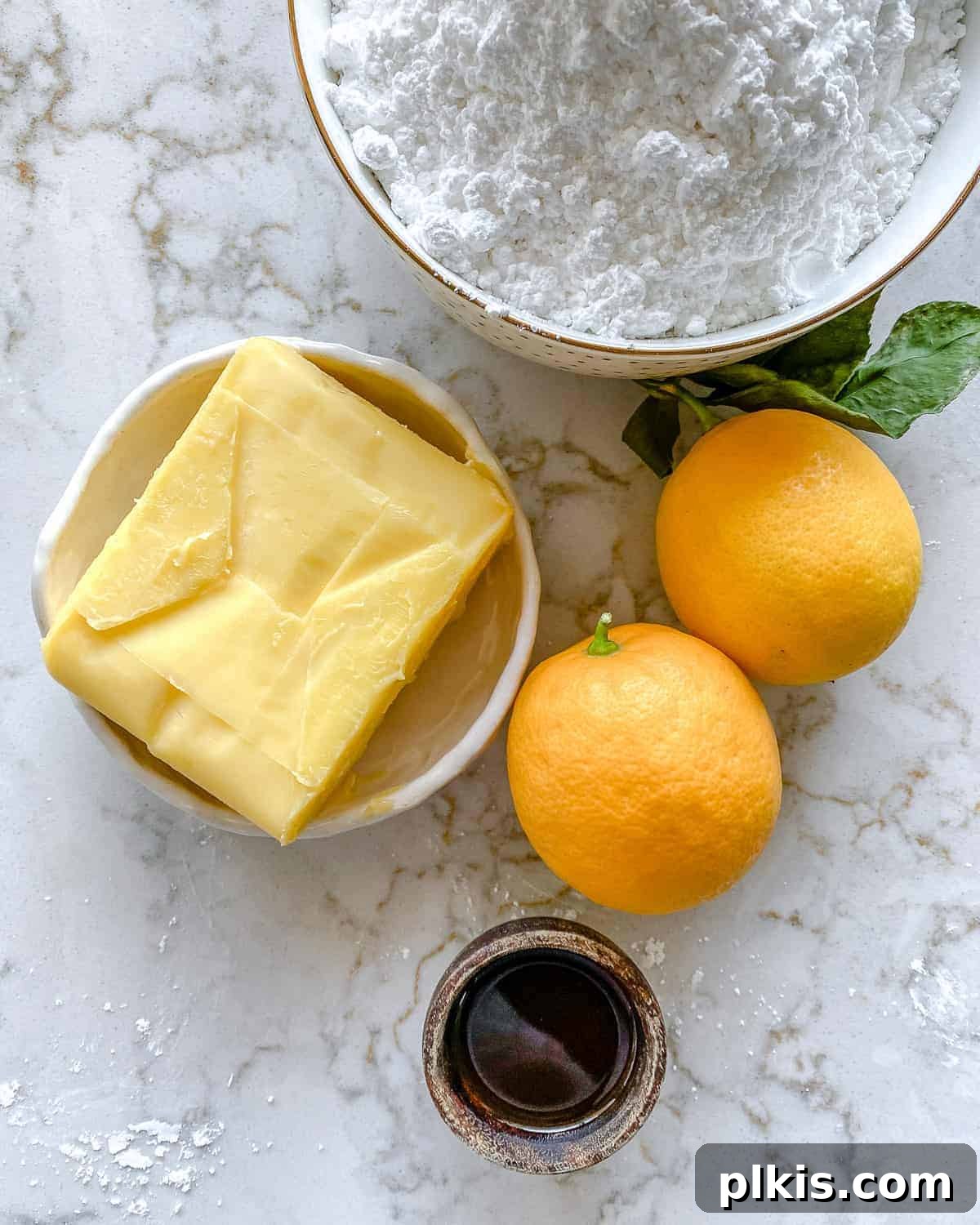 Luscious Vegan Lemon Buttercream 3 A collection of fresh ingredients laid out on a white marble background, including bright yellow lemons, a bowl of white powdered sugar, and a stick of vegan butter, all ready for making vegan lemon buttercream frosting.