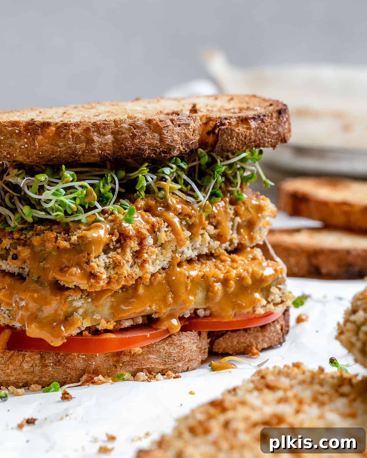 A completed Easy Crispy Buffalo Tofu Sandwich displayed against a clean white background, with a blurred arrangement of ingredients visible in the soft focus background. The sandwich showcases a golden, crispy tofu patty, vibrant red tomato slices, and fresh green sprouts nestled between two slices of whole wheat bread, all generously coated in a rich, orange buffalo sauce.