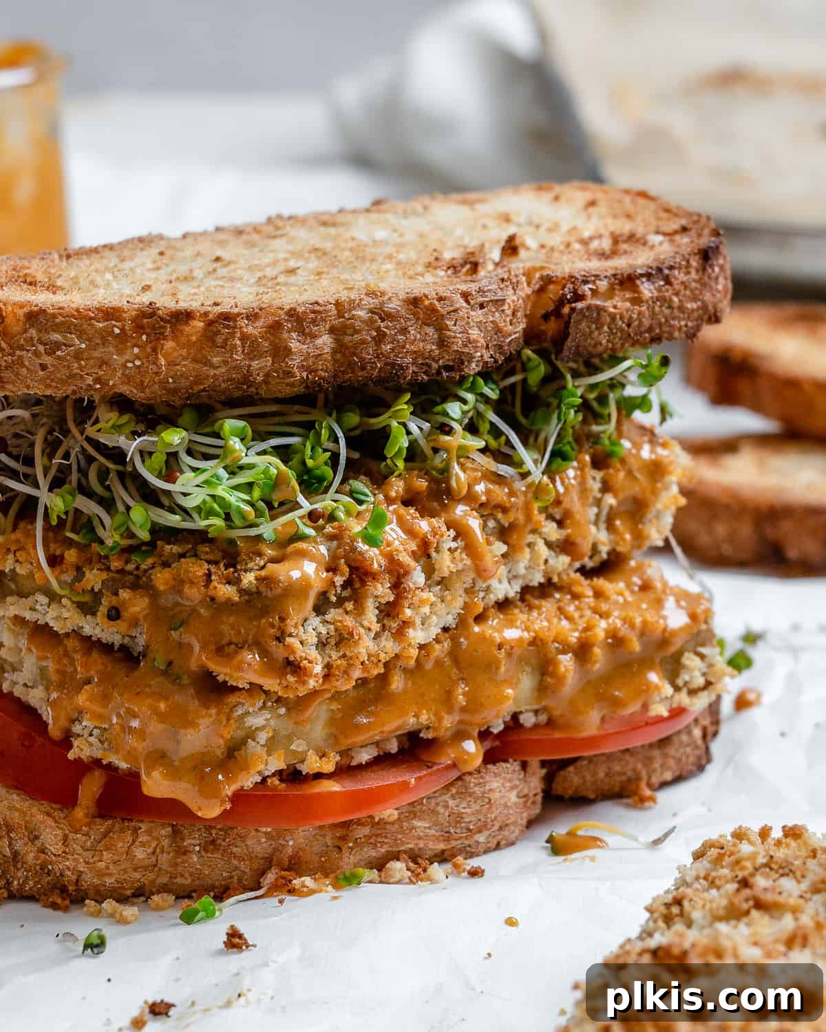 The final, beautifully assembled Easy Crispy Buffalo Tofu Sandwich, positioned against a clean white background with a subtle, blurred hint of ingredients in the background. The crispy tofu patty, vibrant tomato, and fresh sprouts are showcased, drenched in rich buffalo sauce and ready to be enjoyed.
