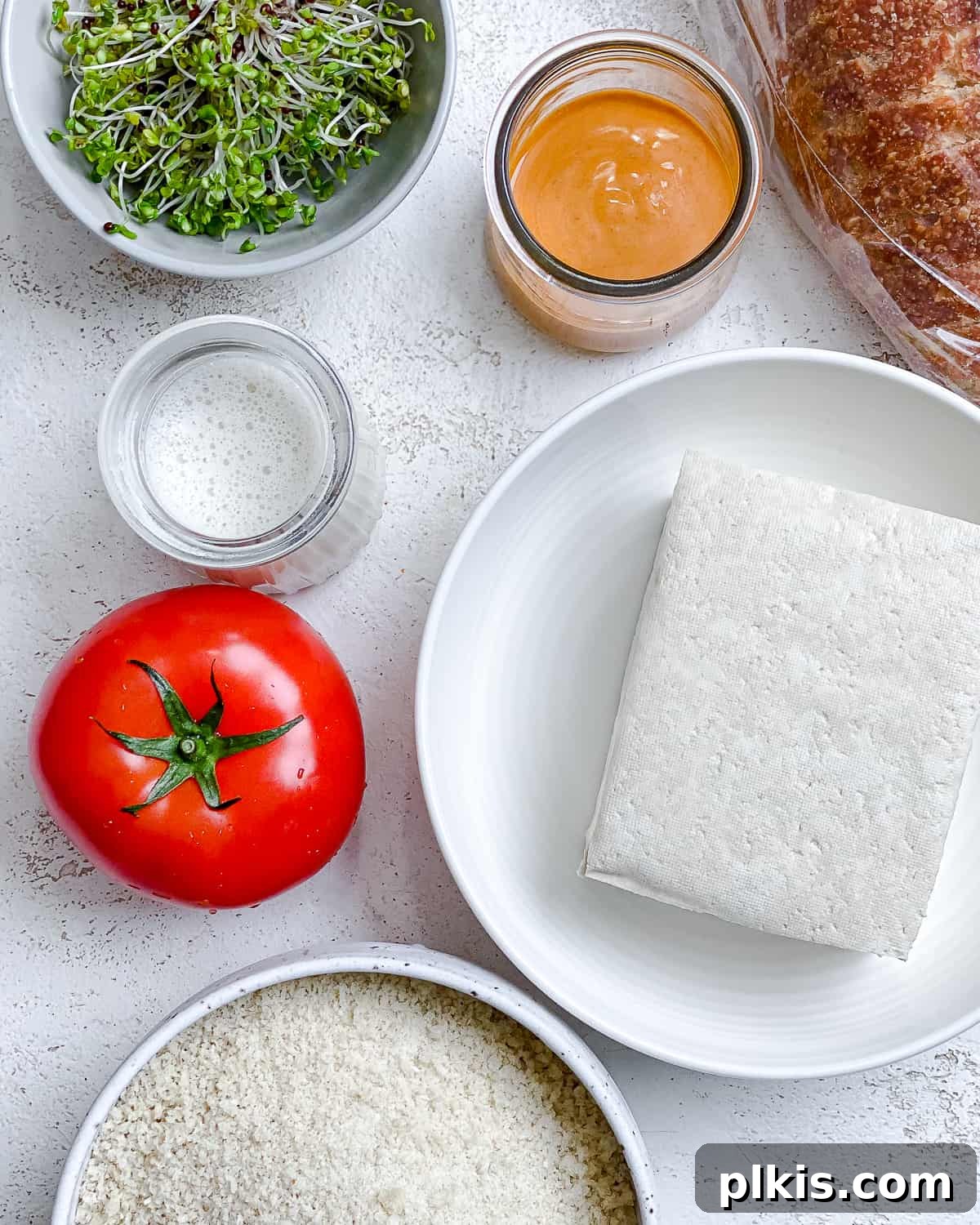 All the pre-measured ingredients for an Easy Crispy Buffalo Tofu Sandwich are neatly arranged against a bright white background, ready for preparation. The arrangement includes a block of tofu, a bowl of plant-based milk, a plate of panko breadcrumbs, a bottle of buffalo sauce, fresh tomato slices, crisp sprouts, and slices of whole wheat bread.