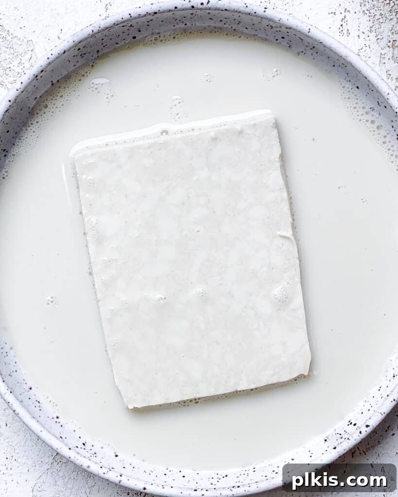 A close-up process shot showing a tofu slice being dipped into a bowl of plant-based milk, part of the breading sequence for the Easy Crispy Buffalo Tofu Sandwich.