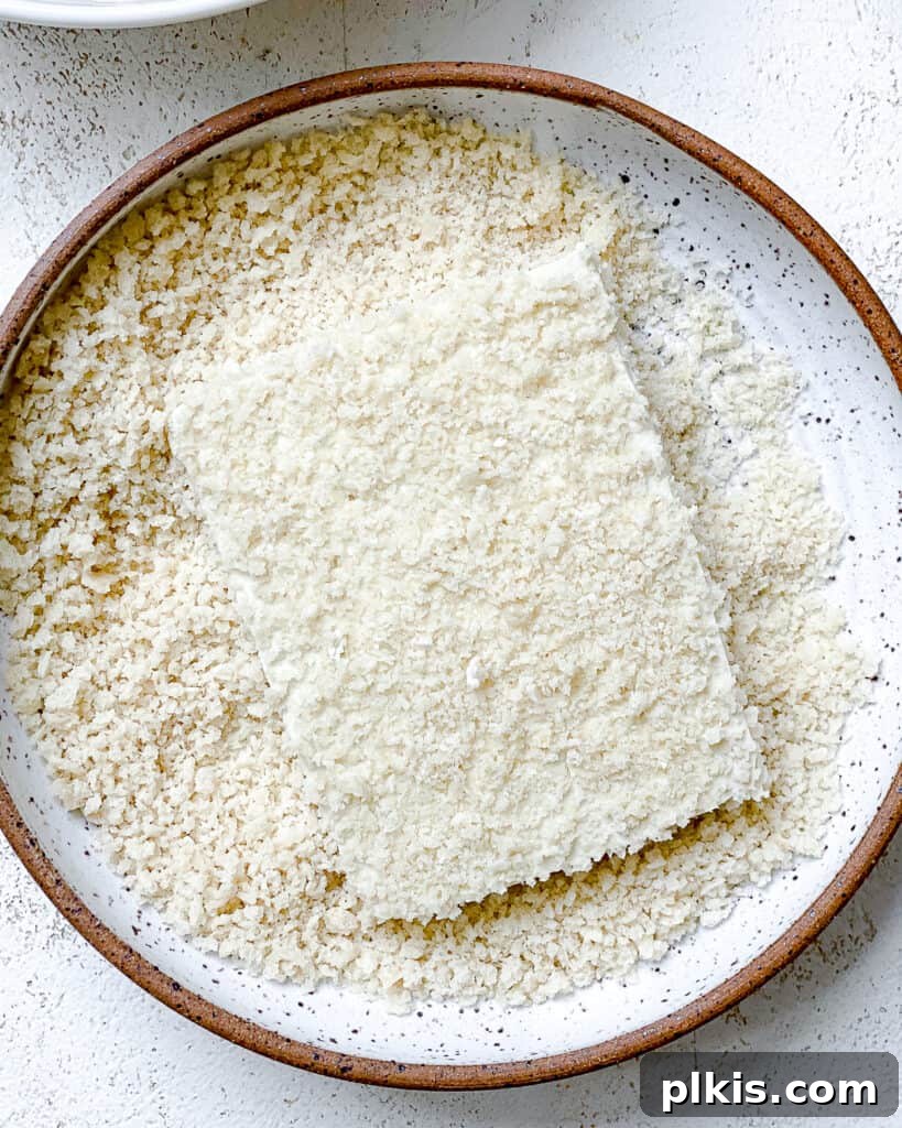 A process shot illustrating a tofu slice being coated in panko-style bread crumbs on a plate, demonstrating the dry stage of the breading for the Easy Crispy Buffalo Tofu Sandwich.