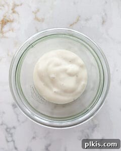 Process of banana cream mixture on top of granola in a glass container against a white marble background