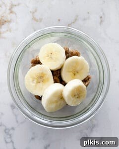 Addition of banana slices on top of granola and whipped cream and banana mixture in clear jar against white marble background