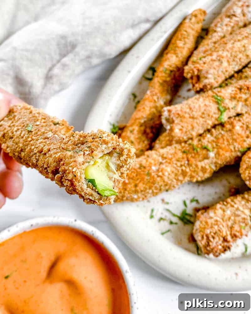 50 Quick Vegan Bites 29 Completed Vegan Baked Zucchini Fries on a white platter, with one fry zoomed in and a small dish of sauce against a white background.