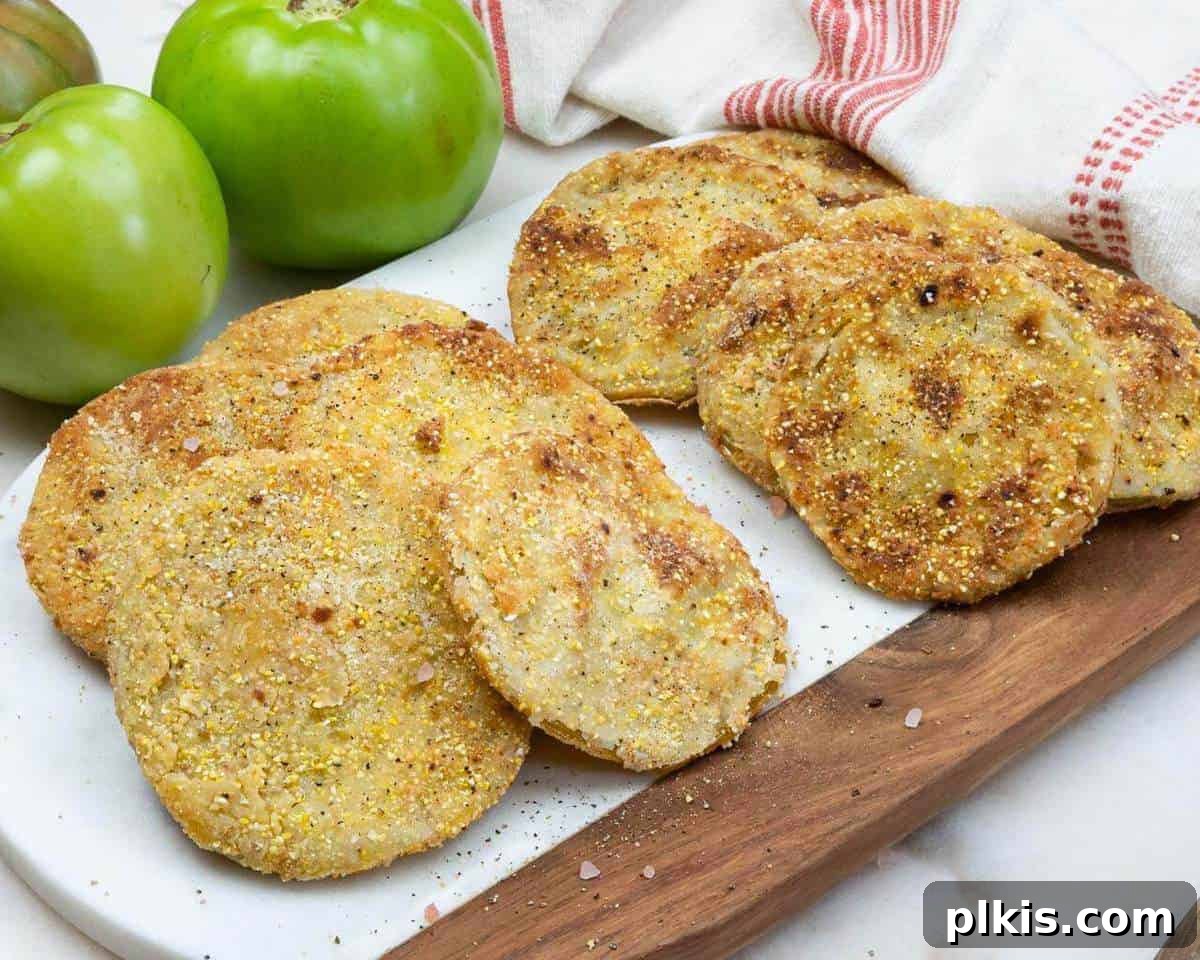 Fried Green Tomatoes Recipe Stack of crispy golden fried green tomatoes on a white background, with fresh whole green tomatoes in the blurred background.