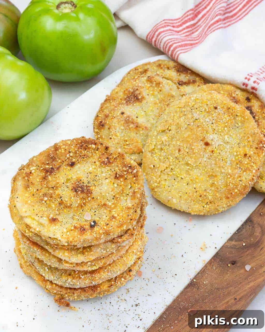 Best Fried Green Tomatoes Close-up shot of a stack of golden brown fried green tomatoes, with more whole green tomatoes blurred in the background, on a white surface.