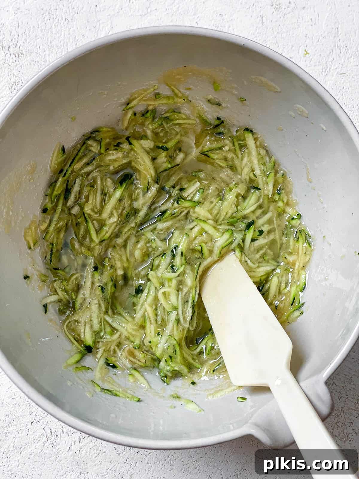 Grated zucchini mixed with wet ingredients in a white bowl, ready for dry ingredients, on a white textured surface.