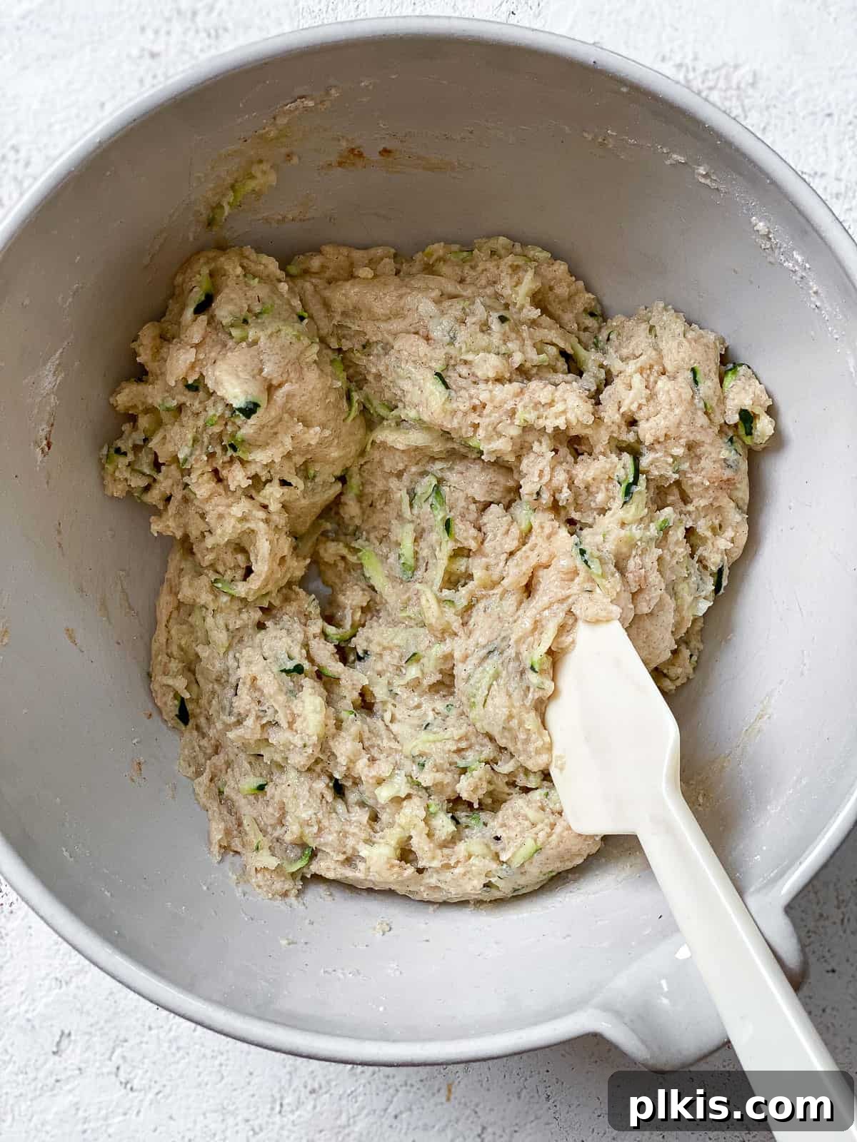 All ingredients for zucchini bread mixed into a thick batter in a white bowl, with a white spatula resting in it, on a white textured surface.