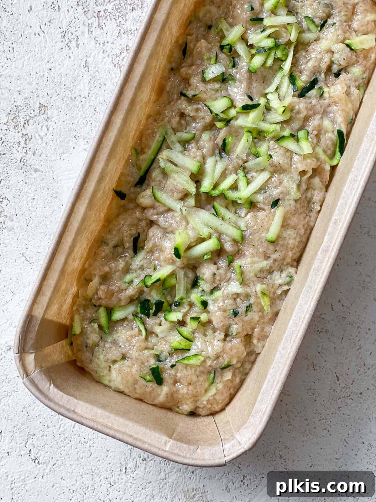 Zucchini bread batter meticulously poured into a greased brown loaf pan, sitting on a white speckled surface, ready for baking.