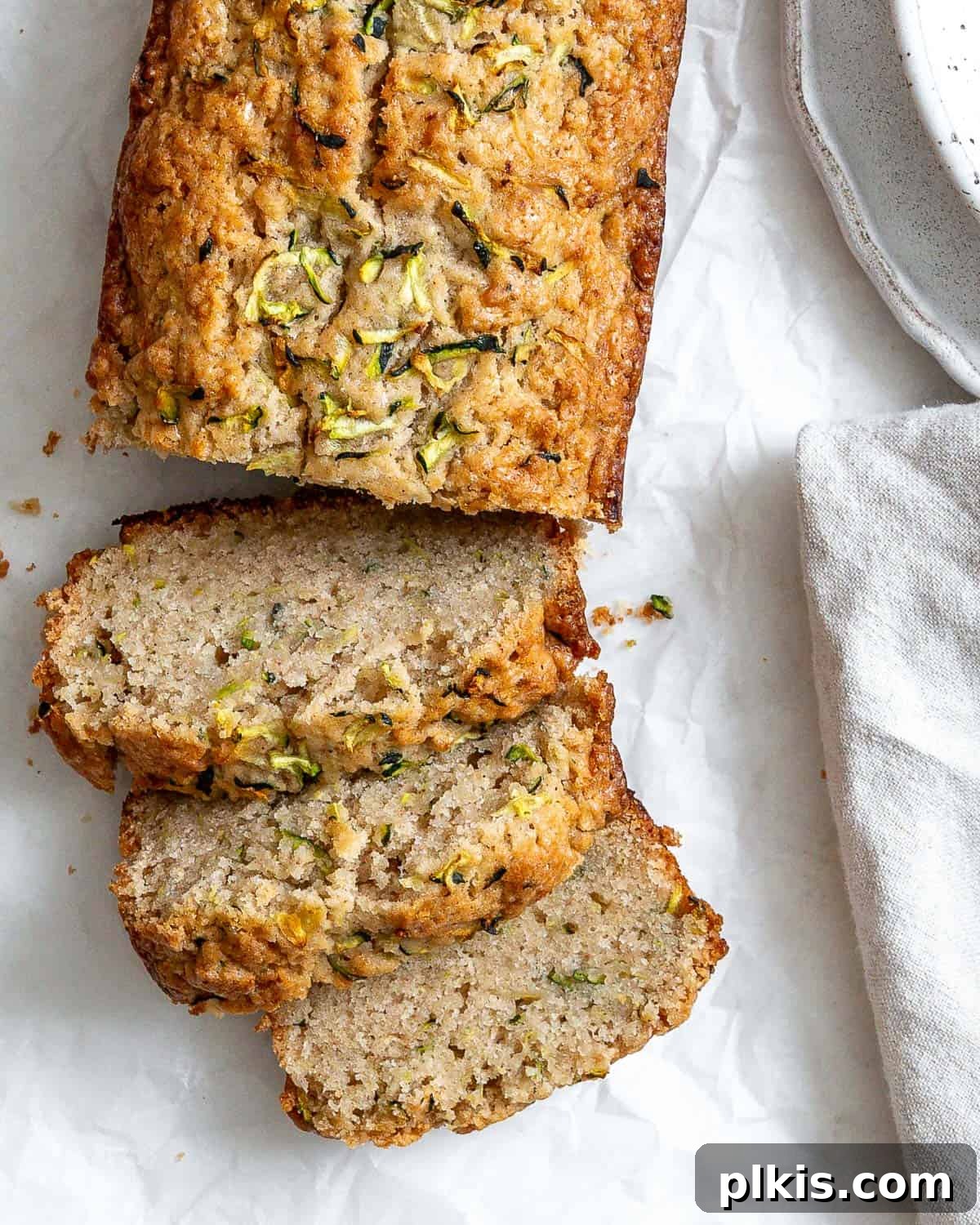 Beautifully sliced vegan zucchini bread on white crinkled parchment paper, with a speckled bowl and gray napkin subtly visible in the background.