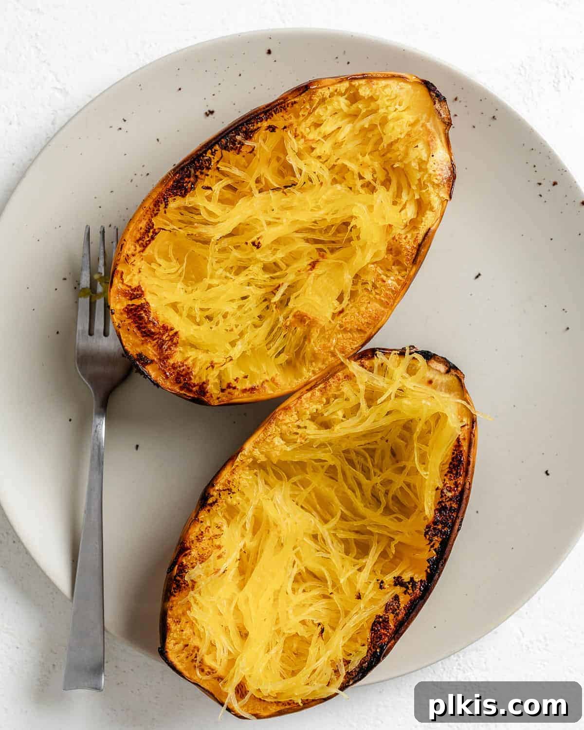 Golden Spaghetti Squash Bowls with Spicy Chili Cheese 6 Cooked spaghetti squash halves resting on a white plate, its tender strands fluffed with a fork.