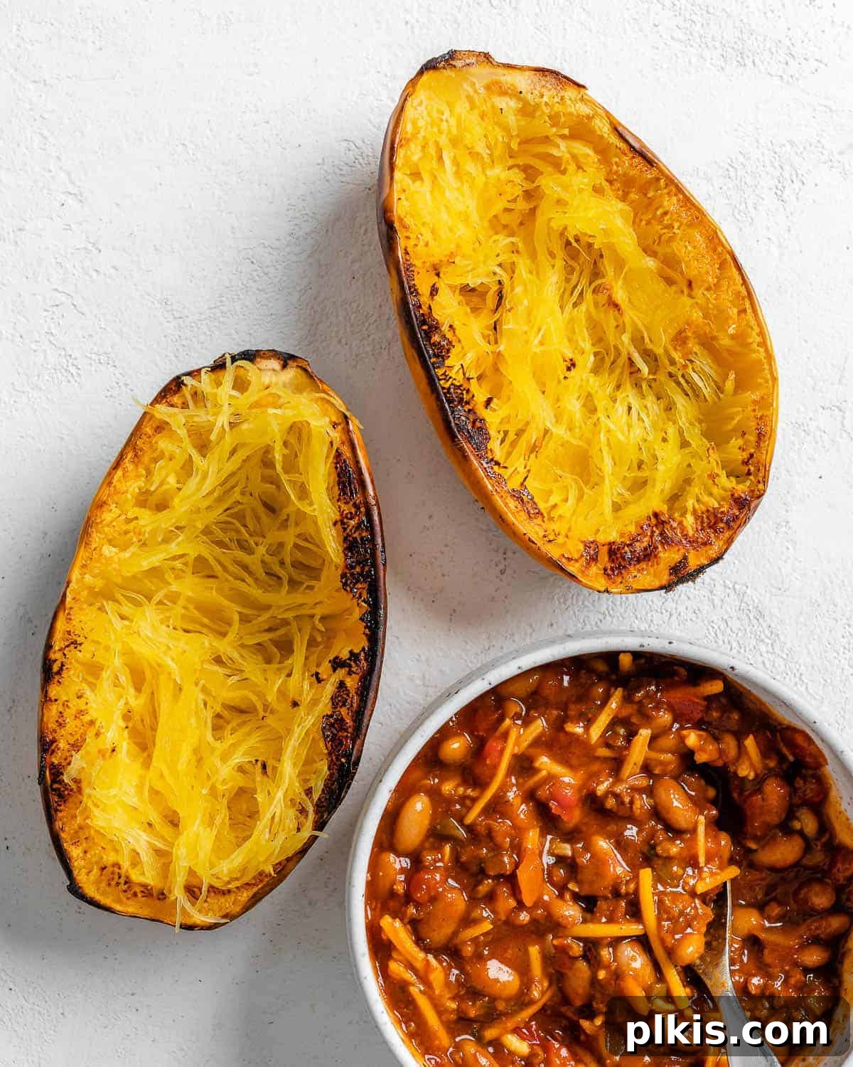 Golden Spaghetti Squash Bowls with Spicy Chili Cheese 8 Two halves of roasted spaghetti squash positioned alongside a bowl of prepared chili on a white background.