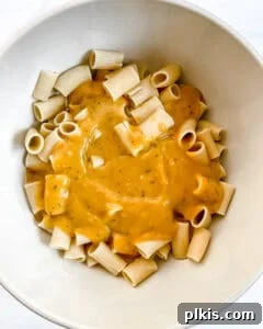 Cooked pasta being tossed with the rich pumpkin sauce in a white mixing bowl, ensuring even coating.