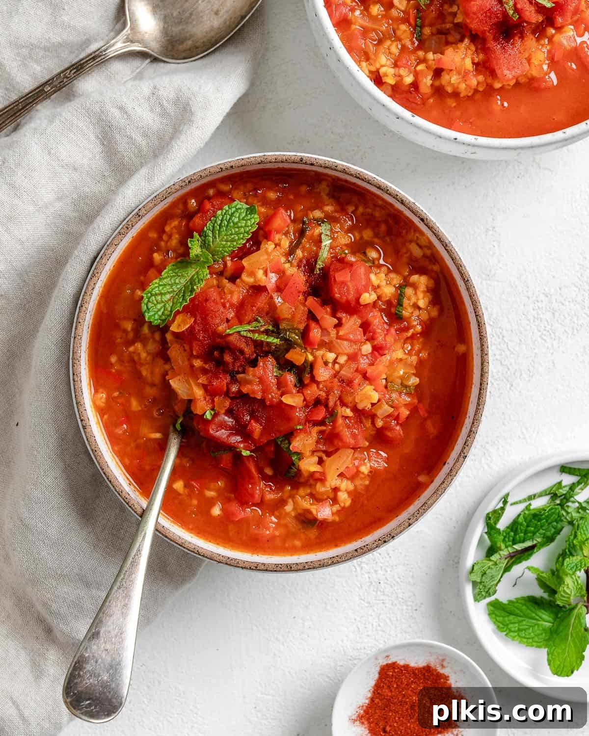 Turkish Embered Pepper Tomato Soup 15 A close-up of a beautifully garnished bowl of Turkish Tomato, Bulgur, and Red Pepper Soup, with fresh herbs and a hint of spice, against a blurred background of extra soup and ingredients.