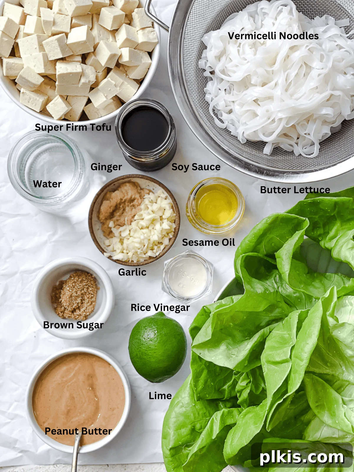 A collection of fresh ingredients laid out on a white surface, including tofu, lettuce, peanuts, lime, and various sauces, ready for making Tofu Lettuce Wraps.