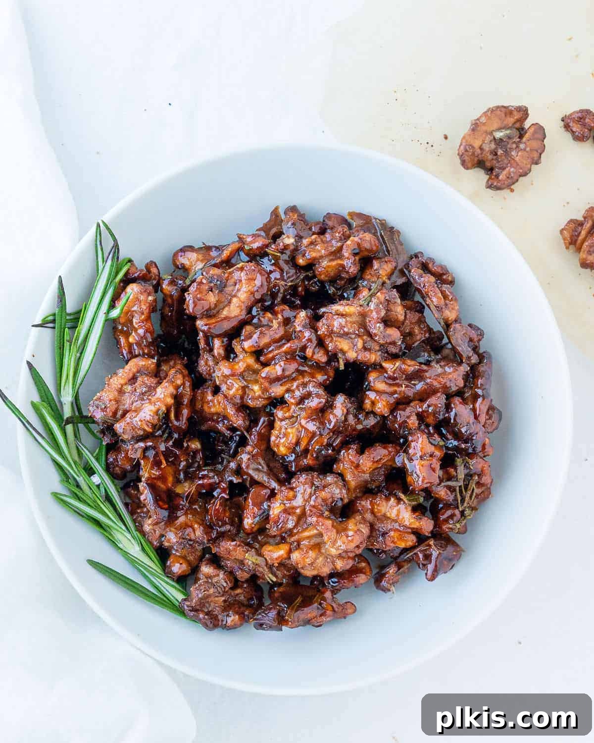 Rosemary Glazed Walnuts 2 Completed sweet rosemary walnuts plated on a white plate against a white surface, ready to be served as a delicious snack or topping.