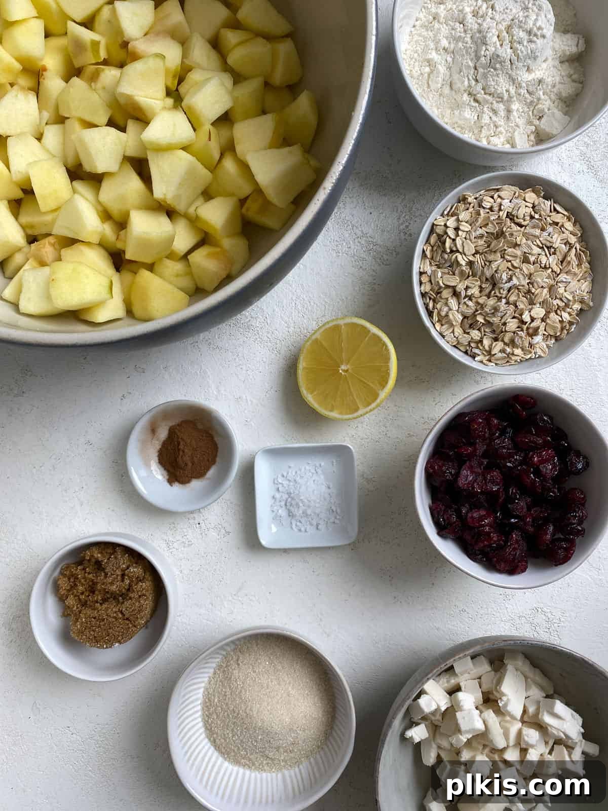 Autumn Apple Cranberry Delight 3 Carefully measured ingredients for Apple Cranberry Crisp, including flour, sugars, vegan butter, rolled oats, lemon juice, cinnamon, dried cranberries, and chopped apples, artfully arranged on a clean white surface.
